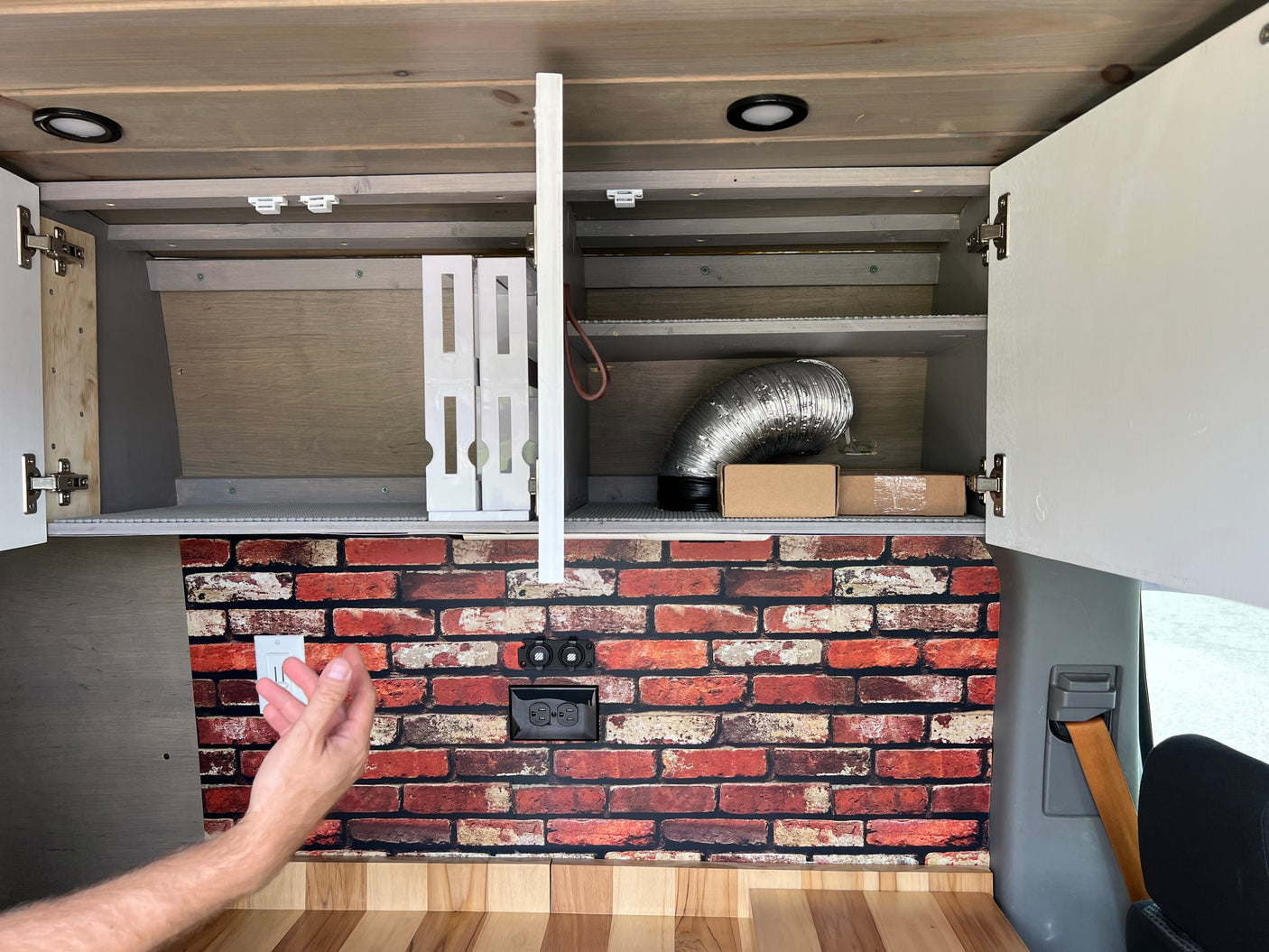 Inside this rv rental, open overhead cabinets reveal storage. A brick backsplash with outlets sits above a wooden countertop.