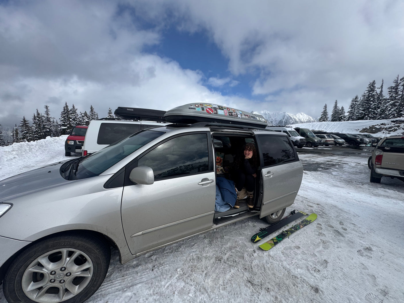 Ski trip stop! A person in a minivan, resembling an RV rental, with skis on snowy ground and mountains in back.