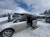 Ski trip stop! A person in a minivan, resembling an RV rental, with skis on snowy ground and mountains in back.