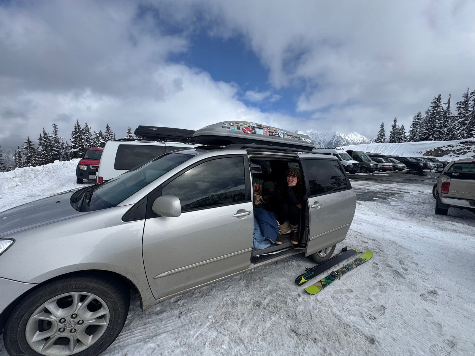 Ski trip stop! A person in a minivan, resembling an RV rental, with skis on snowy ground and mountains in back.