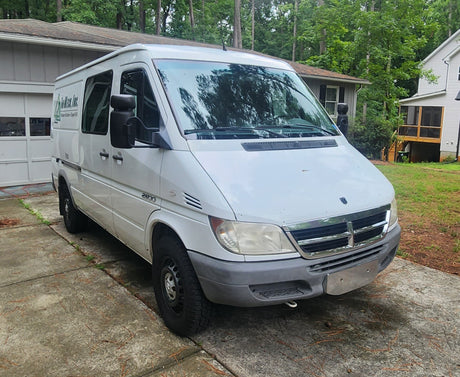 White Dodge Sprinter van on a driveway. Used for cargo or an RV rental. Residential setting with trees.