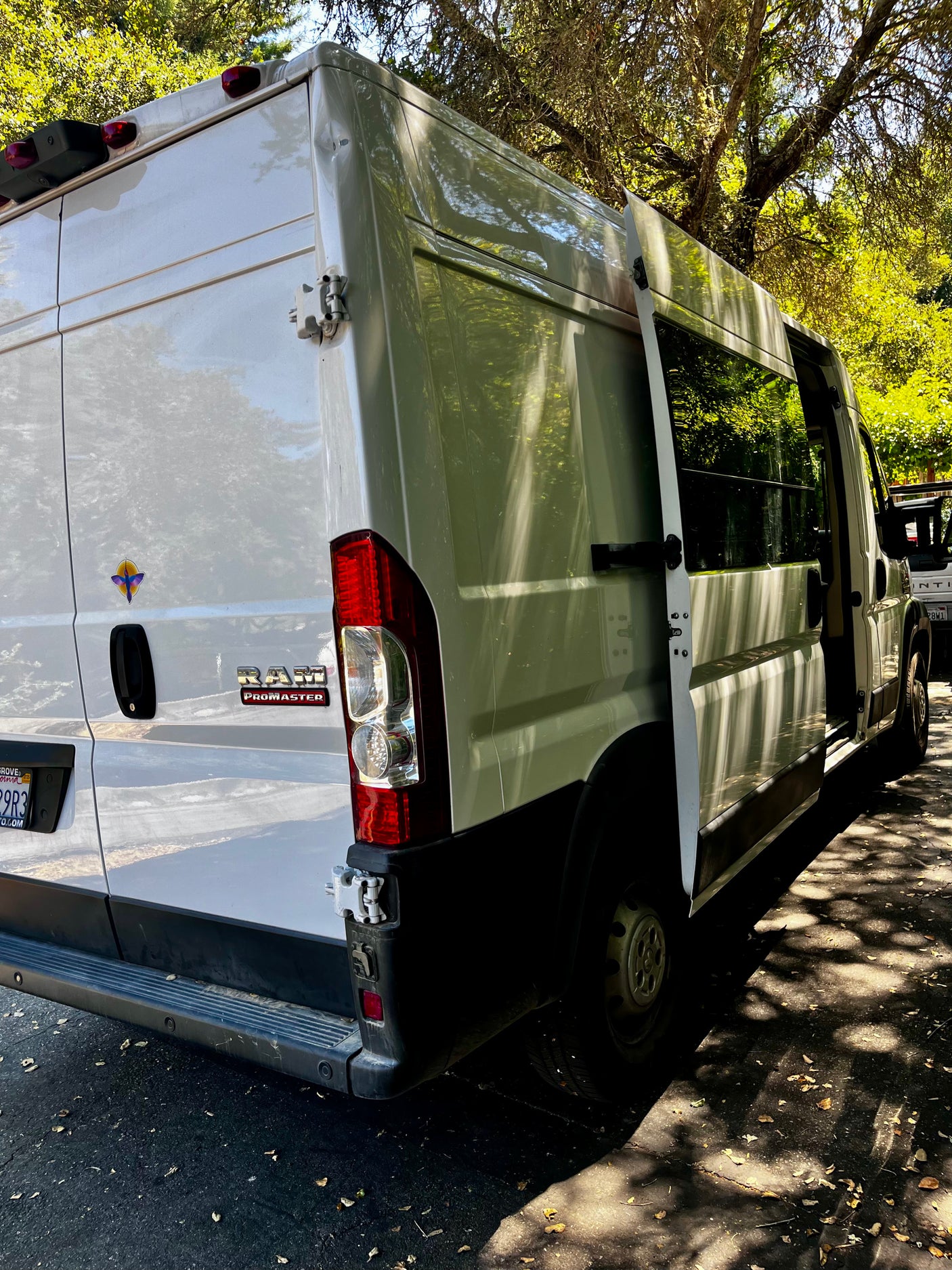 White Ram ProMaster van, configured for RV rental, with its rear and side doors open. Parked outdoors, dappled sunlight on the vehicle and ground.