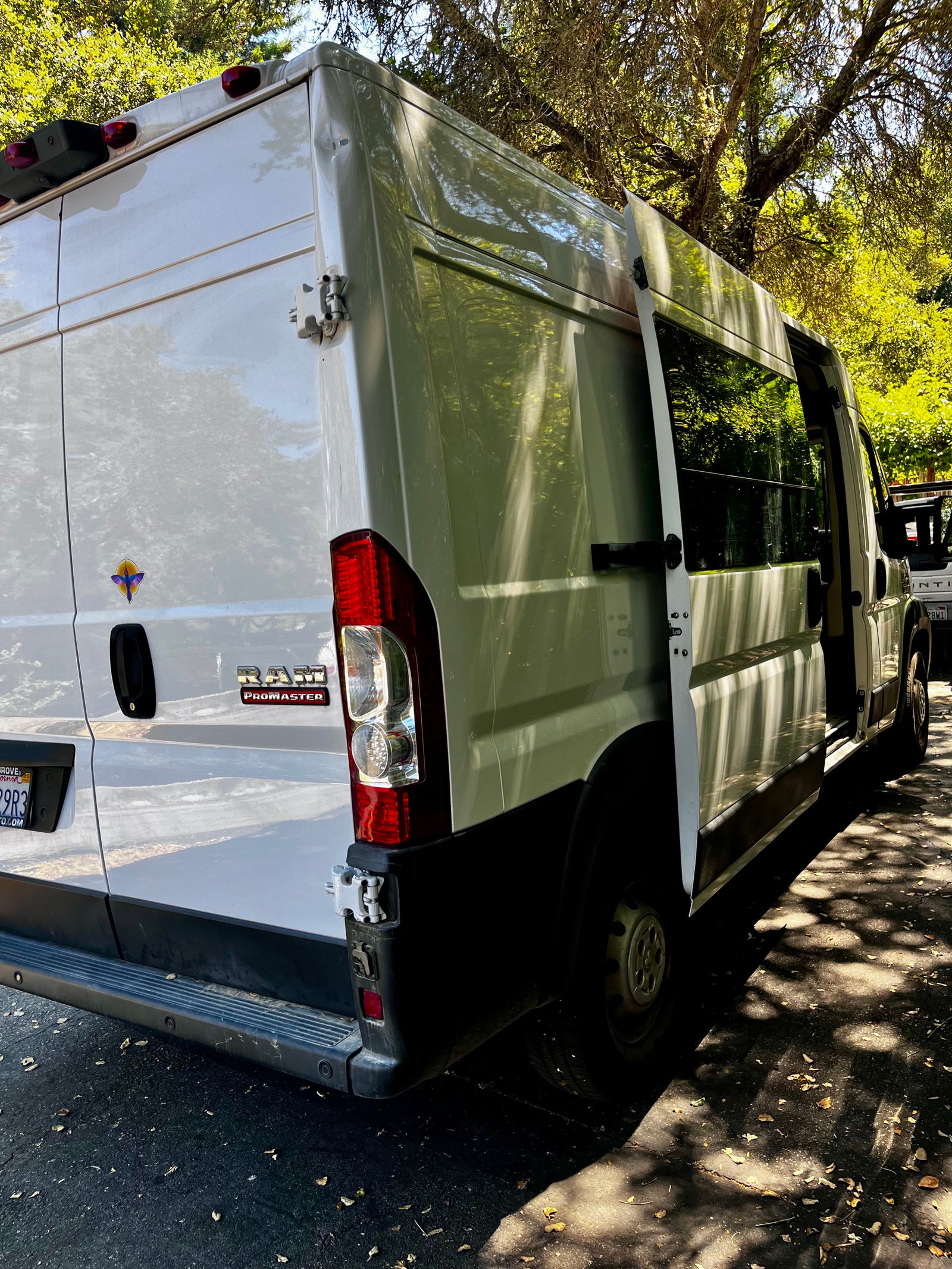 White Ram ProMaster van, configured for RV rental, with its rear and side doors open. Parked outdoors, dappled sunlight on the vehicle and ground.