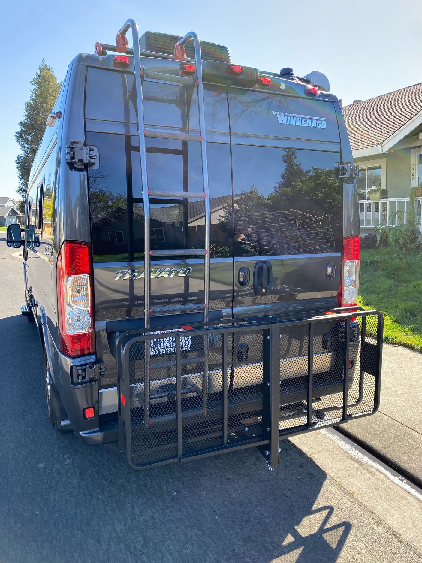 A gray Winnebago Travato RV rental, parked on a street, featuring a rear ladder and a sturdy cargo carrier.