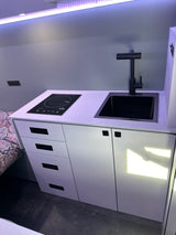 Inside this RV rental, find a modern, compact kitchen with black sink, faucet, induction cooktop, and white cabinets.