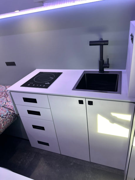 Inside this RV rental, find a modern, compact kitchen with black sink, faucet, induction cooktop, and white cabinets.