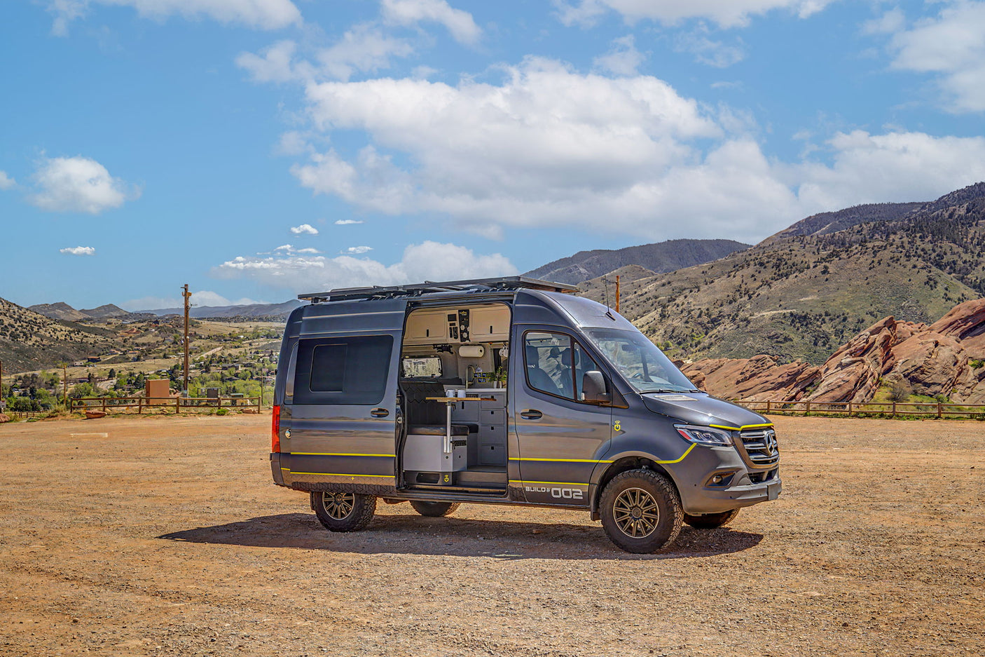 A dark gray custom camper van, "BUILD #002," with open interior, parked on a dirt lot with mountains. Ideal for an rv rental adventure.