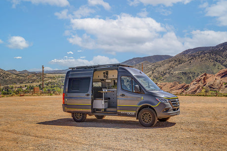 A dark gray custom camper van, "BUILD #002," with open interior, parked on a dirt lot with mountains. Ideal for an rv rental adventure.