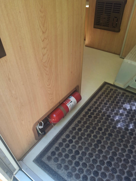RV rental interior: A red fire extinguisher is secured to a light wood wall, beside a textured floor mat.