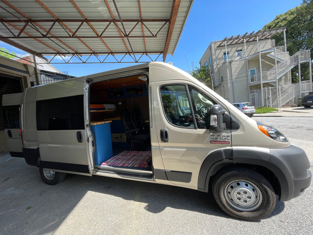 Open side door of a custom Ram ProMaster RV rental, showcasing its unique camper van interior with blue cabinets and wood ceiling.