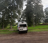 White Winnebago Solis RV rental parked on a dirt path next to a grassy area with tall trees under an overcast sky.