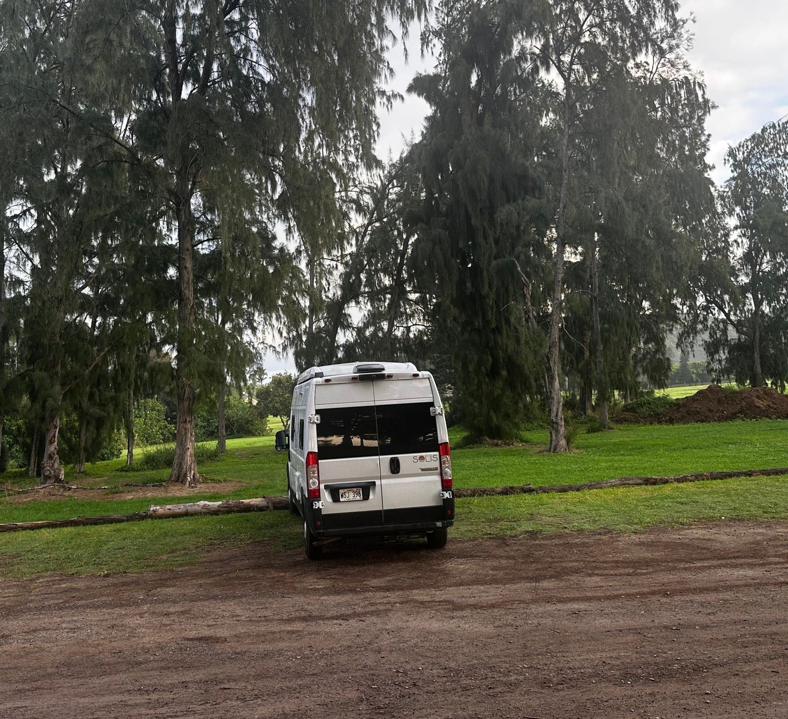 White Winnebago Solis RV rental parked on a dirt path next to a grassy area with tall trees under an overcast sky.