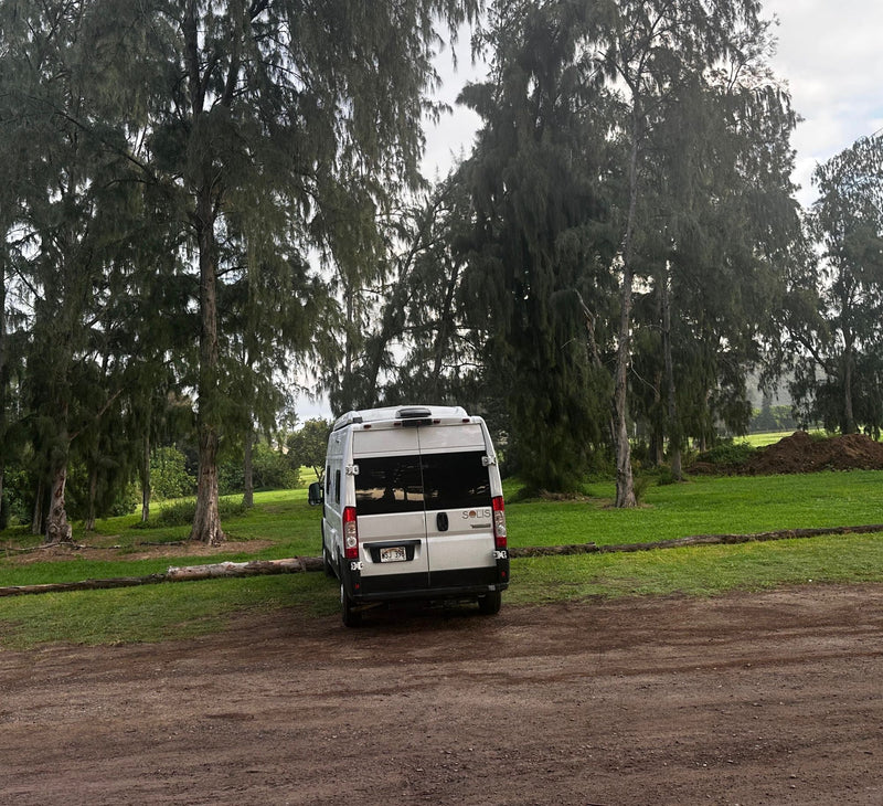 White Winnebago Solis RV rental parked on a dirt path next to a grassy area with tall trees under an overcast sky. — photo 2