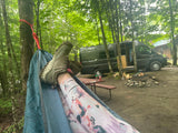 Legs in a hammock at a serene RV rental campsite. A dark camper van, picnic table, and fire pit are visible.
