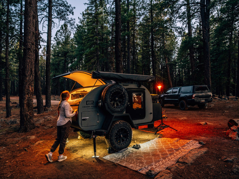 A woman stands by her illuminated Cedar Ridge teardrop camper in a forest at dusk, offering a cozy RV rental camping experience. — photo 17