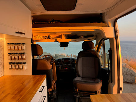 Inside an RV rental van: cozy kitchen with spice rack, front seats, & breathtaking ocean view at golden hour.