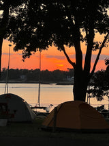 Vibrant sunset illuminates a waterfront campsite. Tents & boat masts silhouetted against the orange sky. Ideal for an RV rental trip.