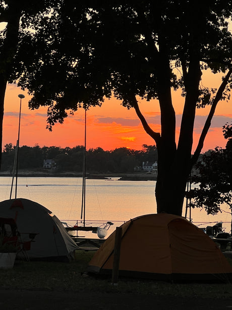 Vibrant sunset illuminates a waterfront campsite. Tents & boat masts silhouetted against the orange sky. Ideal for an RV rental trip.