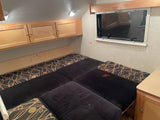 RV rental: Compact interior with a multi-cushion bed, overhead wood cabinets, and an exit window.
