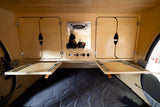 Teardrop RV rental interior: wooden cabinets, control panel with 12V/USB, pull-out tables, and photographer's mirror reflection.