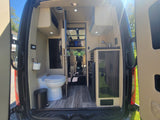 Clean rv rental interior from rear: features a modern toilet, kitchen sink area, and stylish wood flooring.