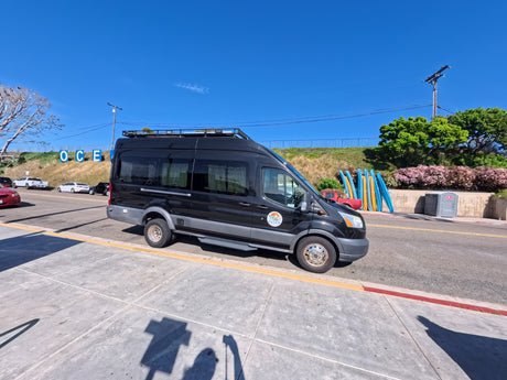 Black **RV rental** camper van parked near an "OCE" sign & surfboards, perfect for **beach vacations** & **travel**.