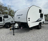 Two Bushwhacker travel trailers, a white "Plus" model in front, are parked on gravel, ideal for an RV rental adventure.