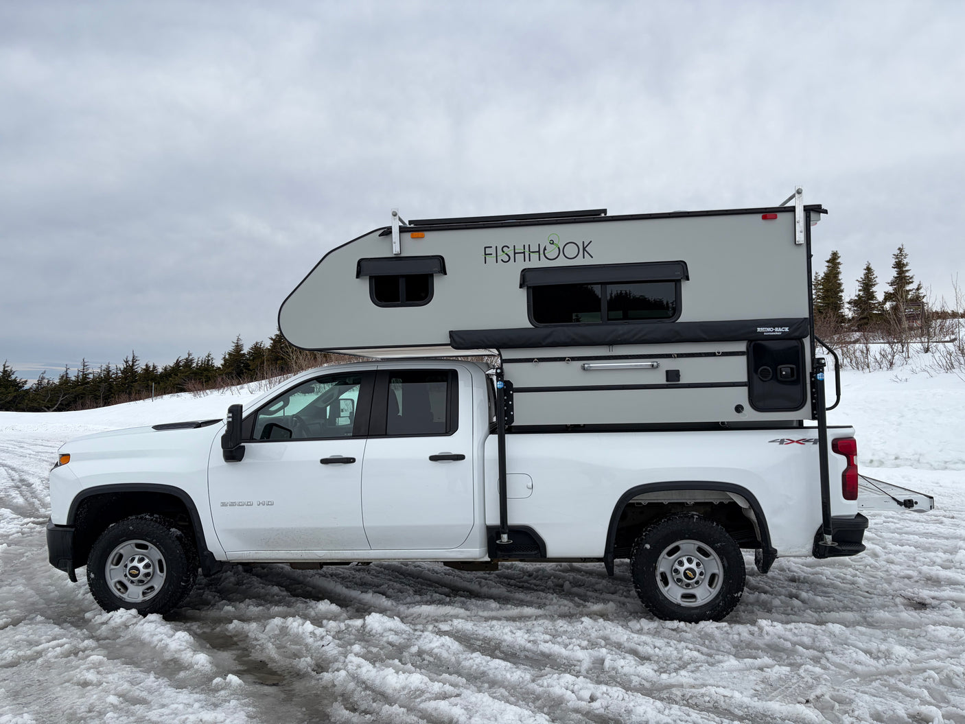 White Chevrolet 2500 HD pickup with a 'FISHHOOK' truck camper on snowy ground, great for an RV rental winter adventure.