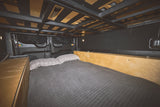 Cozy RV rental bedroom showing a made bed with grey quilted blanket & plaid pillows, light wood cabinetry, and structural overhead storage.