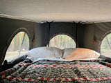 Cozy interior of an RV rental rooftop tent with a comfy bed, patterned blanket, and nature-themed pillows, overlooking a forest.