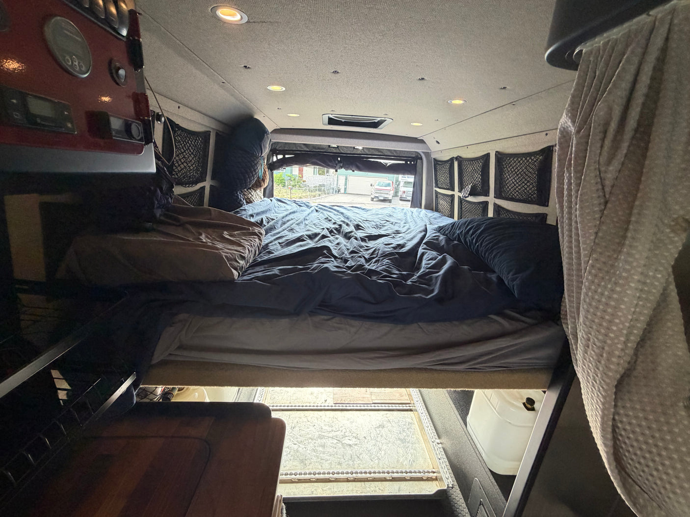Inside a comfy RV rental, featuring a van bed with blue sheets, overhead lights, and practical side storage nets.