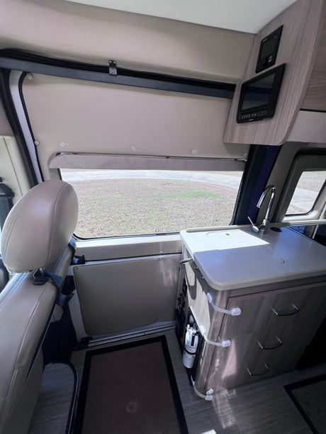 View inside an rv rental, showing a kitchen counter with sink, a headrest, window with grass view, and control panels in a camper van.