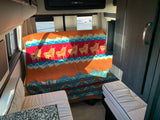 RV rental interior with a cozy bed covered by a vibrant deer-patterned blanket, light seating, and dark cabinetry.