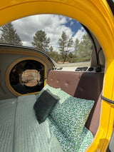 Explore nature with this RV rental! Cozy interior shows a bed, round porthole, & scenic pine tree views from large windows.