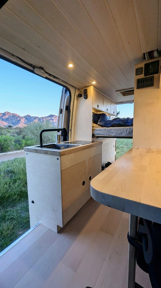 Modern RV rental interior with kitchen, sink, and bed, featuring wood accents and scenic mountain views.