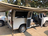 White van, possibly an RV rental, with an extended side awning and open doors, revealing a basic conversion interior for camping trips.