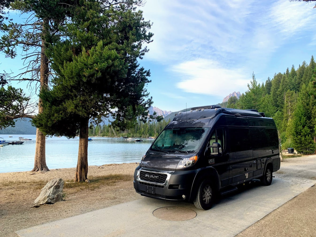 This black RV rental is parked by a tranquil lake with boats, flanked by tall trees and mountains. Ideal for scenic adventures.