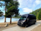 This black RV rental is parked by a tranquil lake with boats, flanked by tall trees and mountains. Ideal for scenic adventures.
