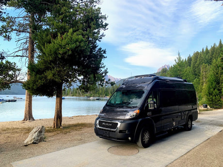 This black RV rental is parked by a tranquil lake with boats, flanked by tall trees and mountains. Ideal for scenic adventures.