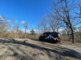 A black RV rental van with a white mountain graphic, parked on a gravel lot, surrounded by bare trees under a blue sky.