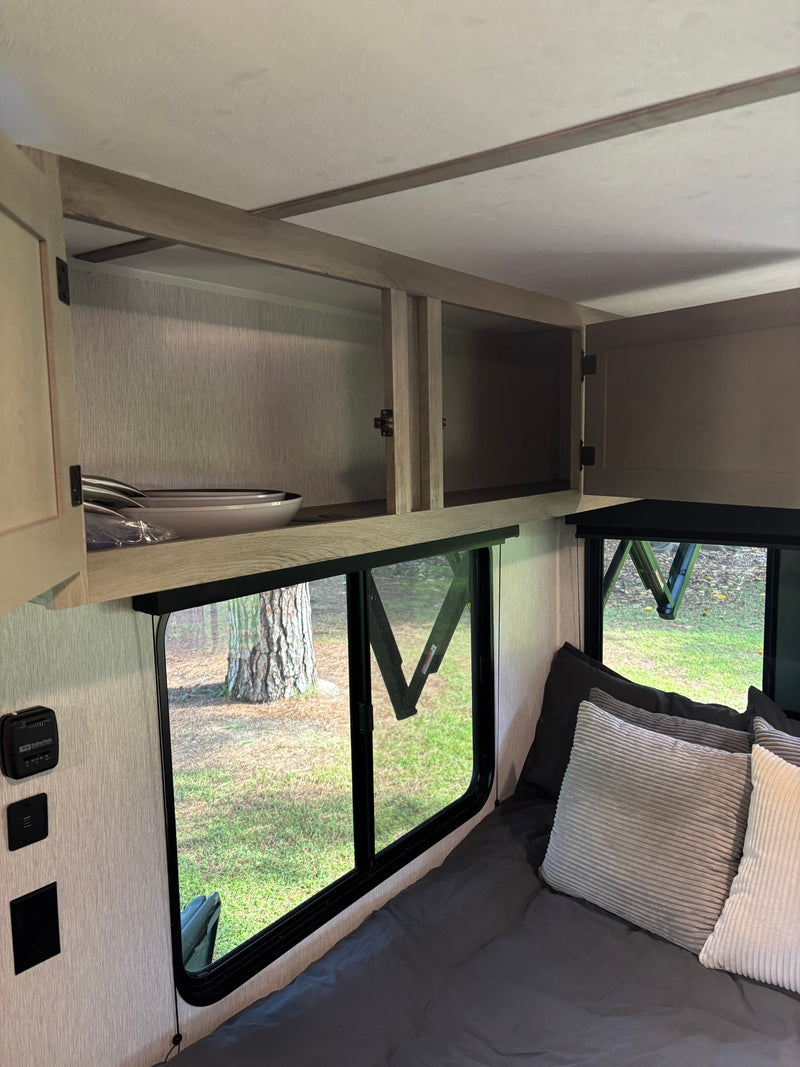 RV rental interior: open overhead cabinets with dishes, large windows overlooking trees, a thermostat, and a made-up bed. — photo 12