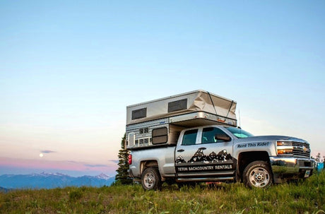 Teton Backcountry Rentals RV rental: A silver pickup truck with a pop-up camper on a scenic mountain hilltop at dusk.