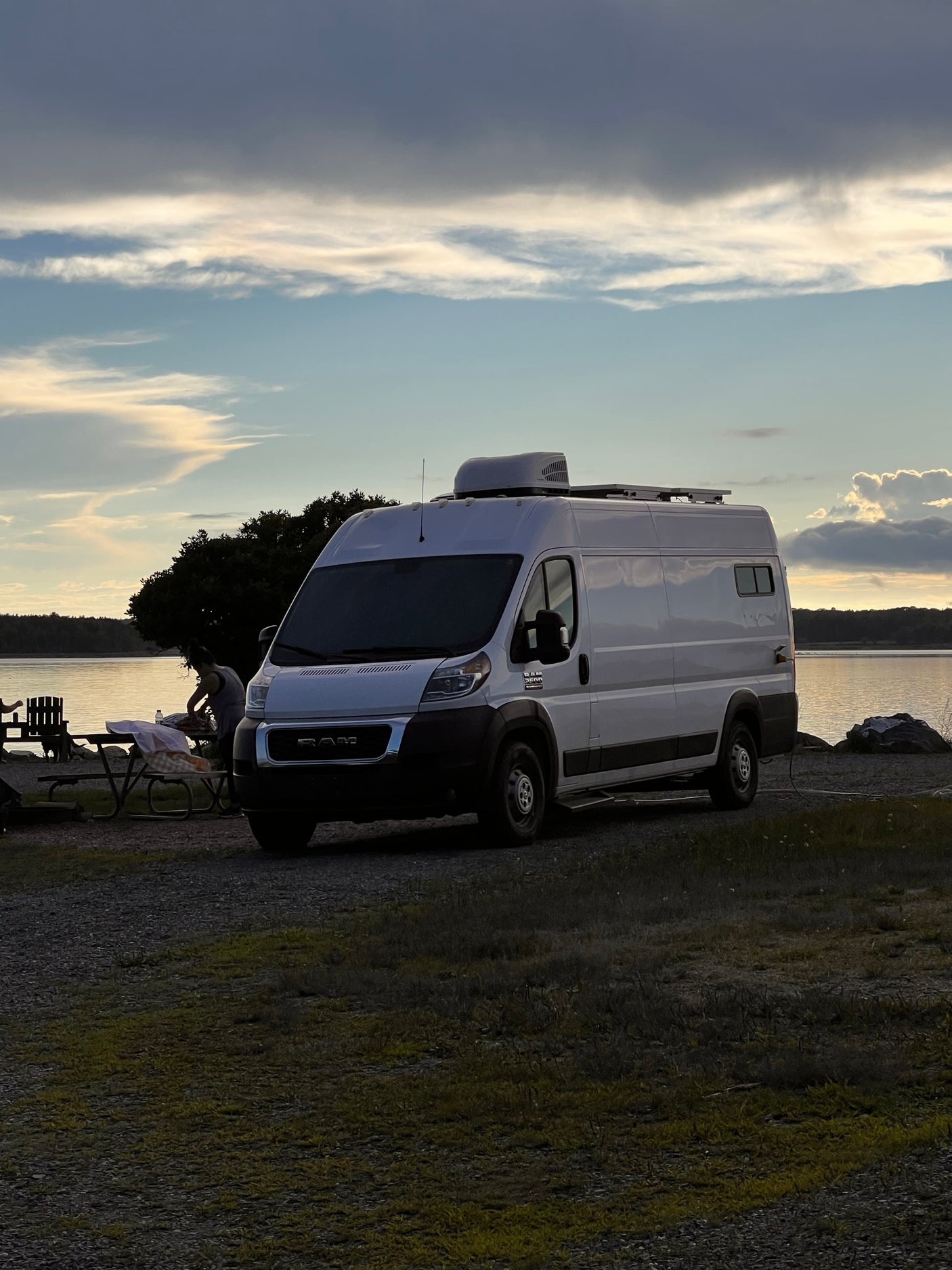 RV rental: White camper van parked at a scenic lakeside spot at dusk, with someone by a picnic table setup.