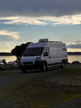 RV rental: White camper van parked at a scenic lakeside spot at dusk, with someone by a picnic table setup.