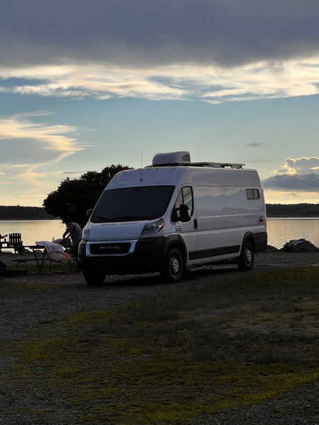 RV rental: White camper van parked at a scenic lakeside spot at dusk, with someone by a picnic table setup.