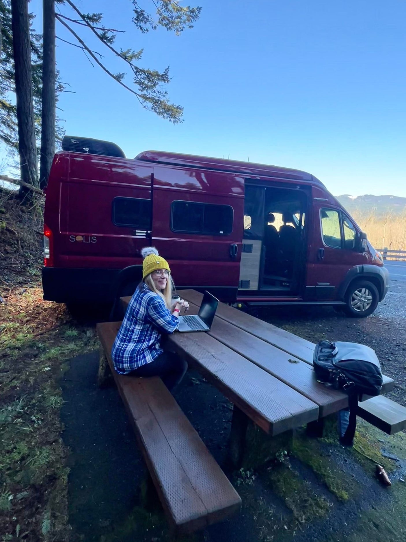 A woman works on a laptop at a picnic table next to her red Solis RV rental van, parked amidst trees.