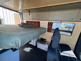 Inside a spacious RV rental camper van, featuring an elevated bed, rear seating, window views, and a portable fridge.