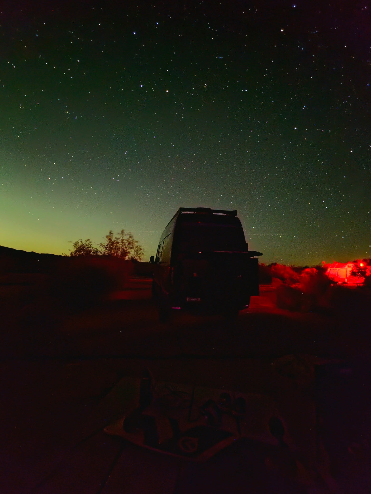 RV rental for stargazing: A dark camper van beneath a vibrant green, starlit sky, with hints of red light in the distance.