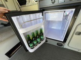 A person's hand holds open a compact fridge in an RV rental, revealing four green Carlsberg beer bottles inside. Great camper amenity.