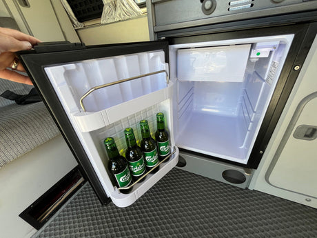 A person's hand holds open a compact fridge in an RV rental, revealing four green Carlsberg beer bottles inside. Great camper amenity.